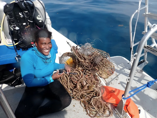 Tokea Payton sits on a boat next to a pile of trash removed from the ocean. She holds a ruler and a small camera.