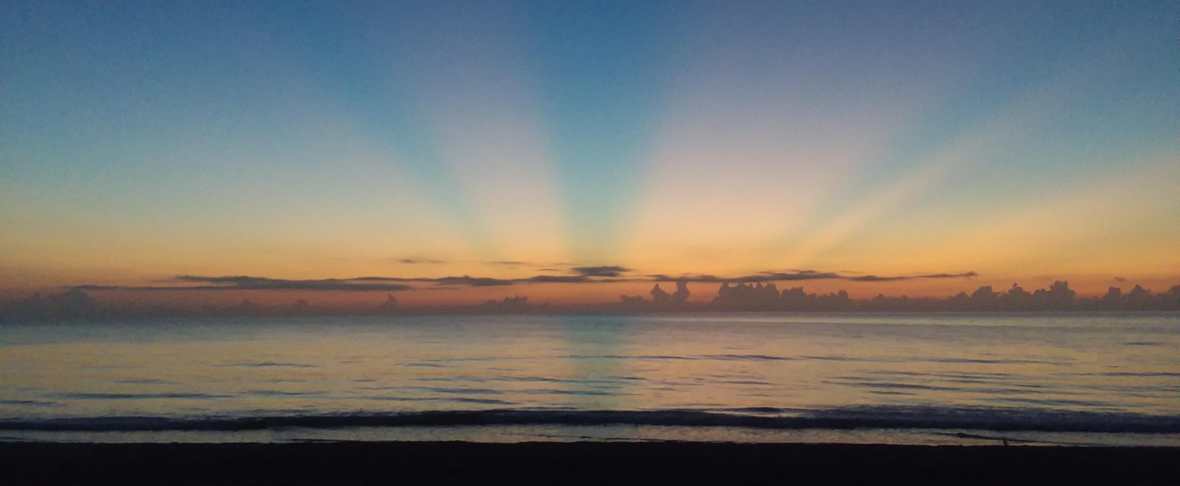 Photo of a colorful sunrise over the horizon of a calm ocean offshore of Melbourne, Florida. The morning light is barely peeking, blending into the night.