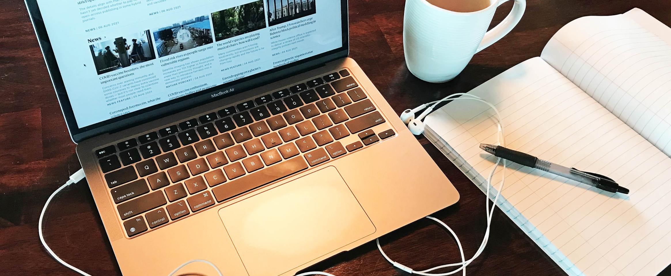 Horizontal photo of a laptop open on a wood grain desk, staged with headphone cords, an open notebook, pen, and mug. Original photo credit Alex Witze.
