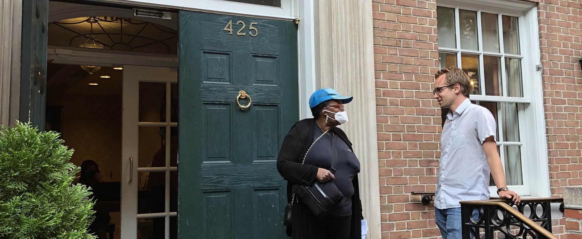 Photo of Neelise, a Fountain House member, talking with Andrew, a Fountain House and N A S W member, on the doorsteps of Fountain House's Manhattan building in New York City. The building has old double doors and a ring knocker, and a stone beam engraved with the organization's name overhead.