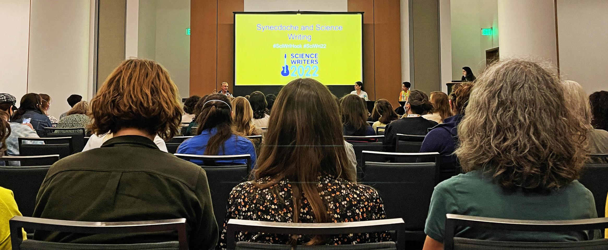 Photo focused on the back of the heads of three audience members amid a sea of attendees in the foreground, while in the background sit panelists David Quarmmen, Sabrina Imbler and Mara Grunbaum below a projector screen with slide title Synecdoche and Science Writing, with moderator Maya L. Kapoor standing at the podium to stage left.