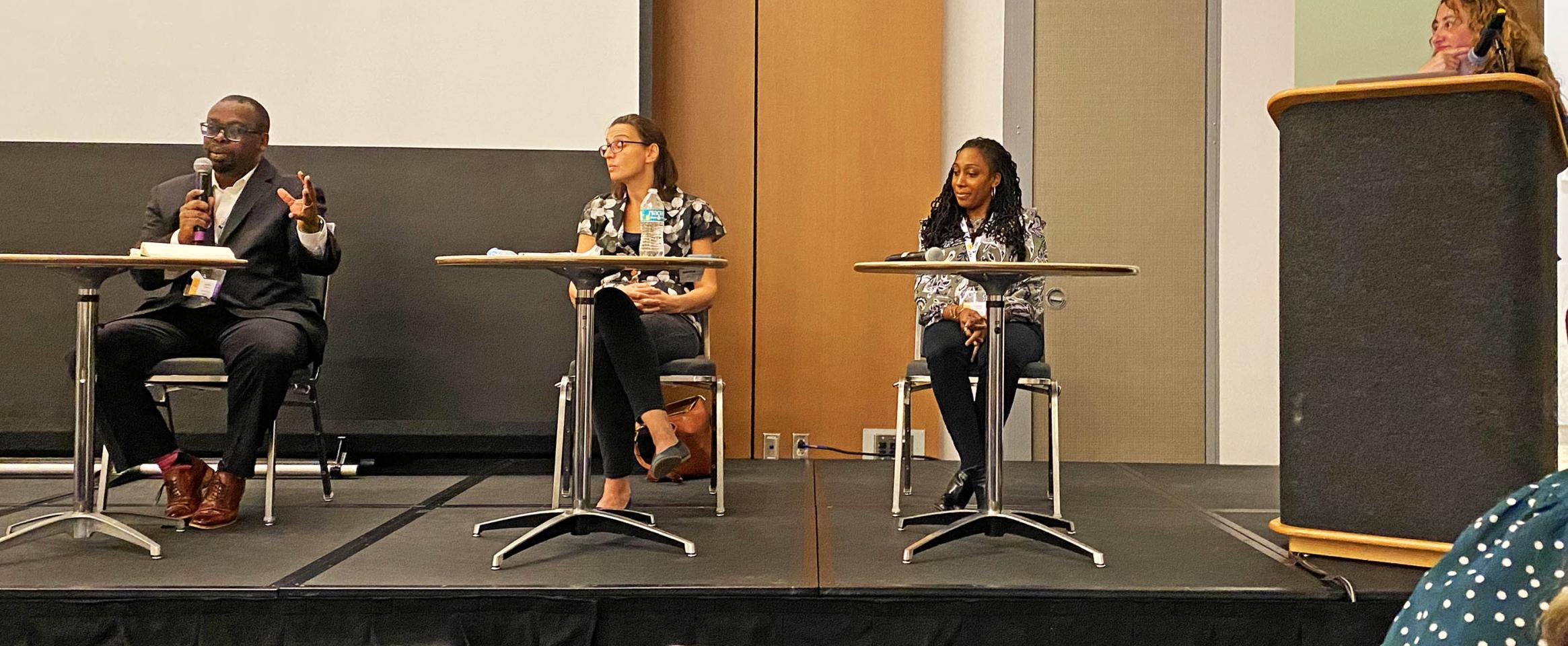 Horizontal photo of Jermey N. A. Matthews, Luba Ostashevsky, and LLathea Mondesir, seated on stage, and moderator Lina Zeldovich, standing at podium.