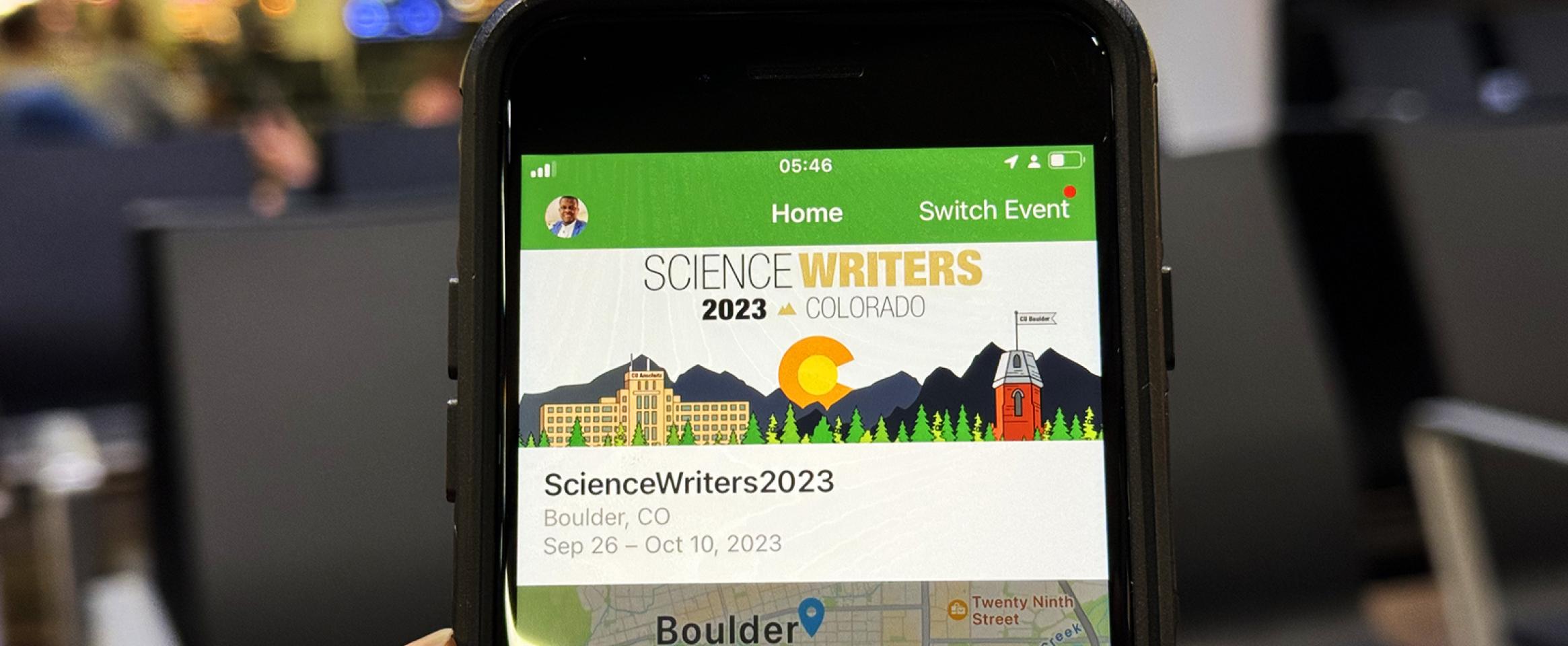 Horizontal photo of a melanated hand holding up a smart phone showing the Science Writers 2023 Whova conference homepage, In the background, airport seating and people are visible. Photo by Paul Adepoju
