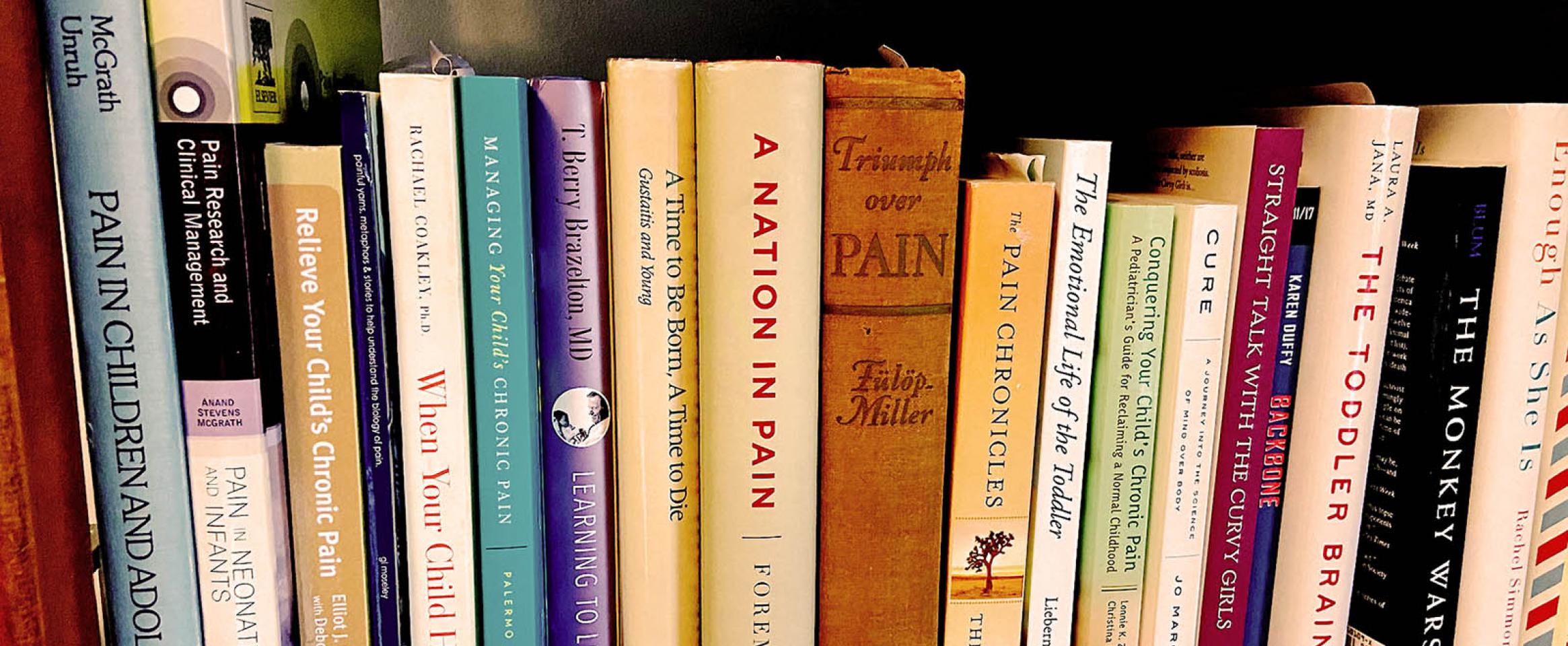 Rectangular photo of a row of books on a wooden bookshelf, with many titles related to the science of pain and children's health, some new and some visibly old.