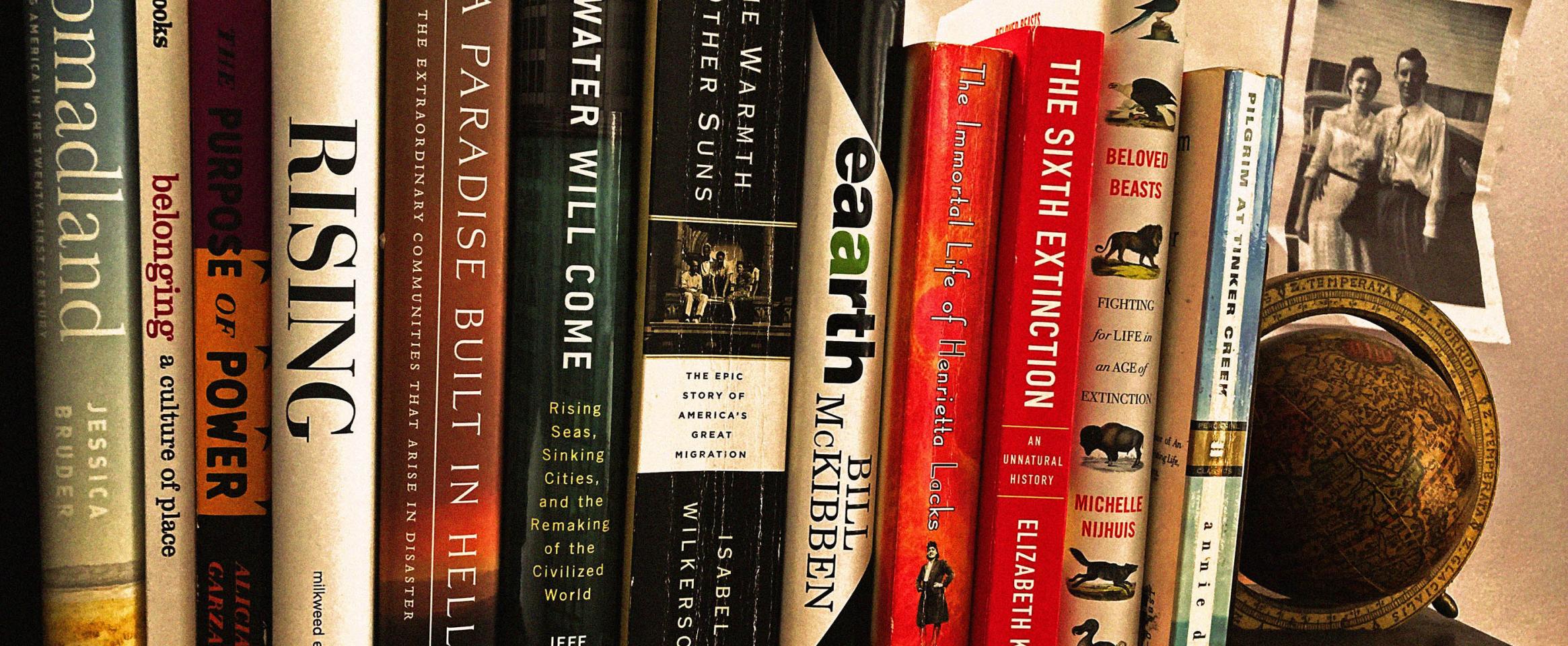 Rectangular photo of a row of books on a wooden bookshelf, with many titles related to the earth, environment, and science. A bookend with an antique globe, and a monochrome vintage photo of a couple are visible adjacent.