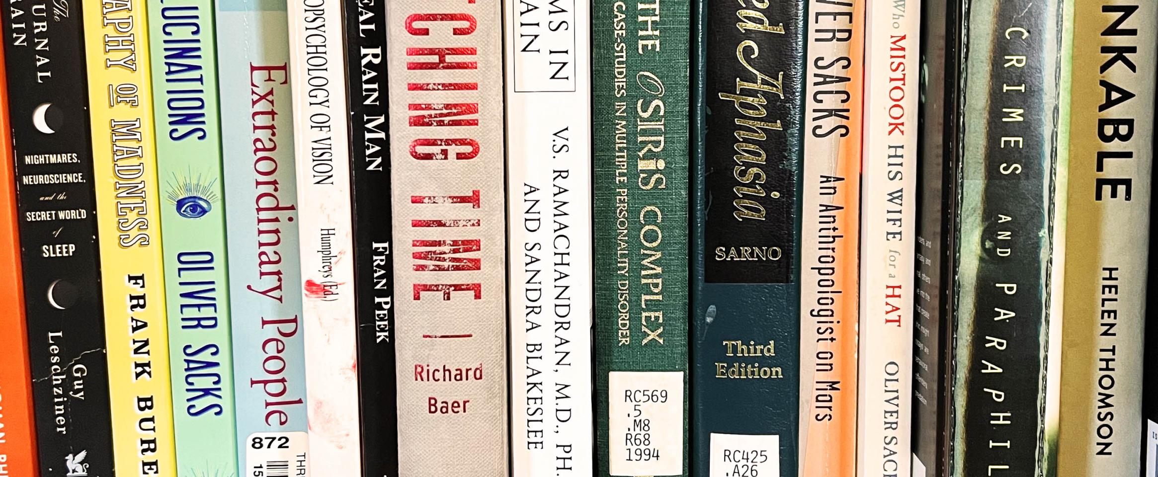 Horizontal photo of a bookshelf with various book spines visible, many with library catalog stickers, and a few authored by Oliver Sacks. Photo by Marc Dingman