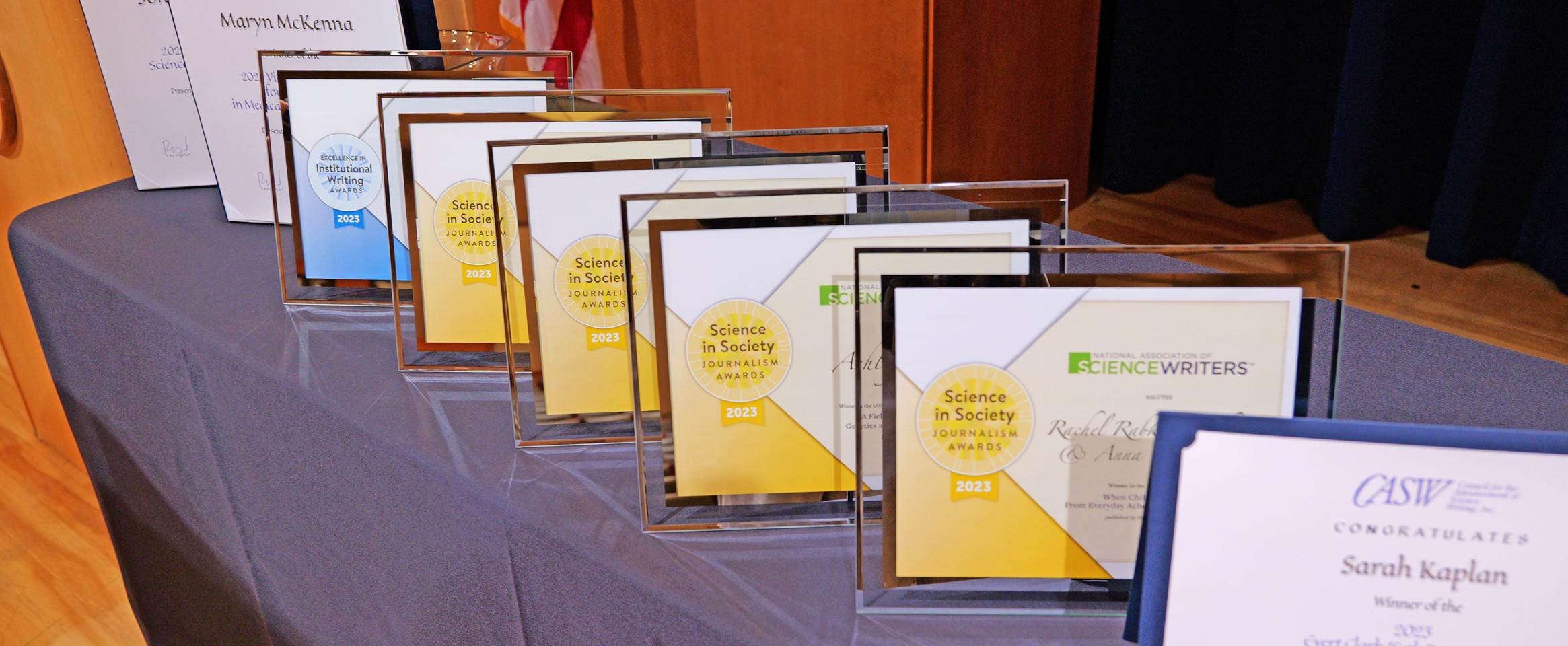 Horizontal photo of glass-framed N A S W awards alongside cardstock C A S W awards on a tableclothed table. Photo by Casey A. Cass University of Colorado Boulder