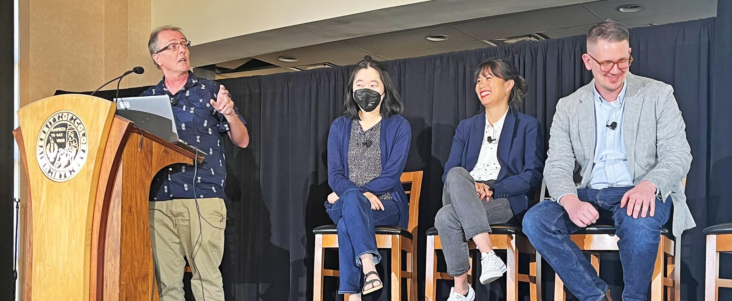 Horizontal photo of Peter Aldhous speaking at the podium next to seated panelists Lisa Song, who is masked, Stephanie Lee, and Nicholas Florko. Photo by Gabriella Lewis.