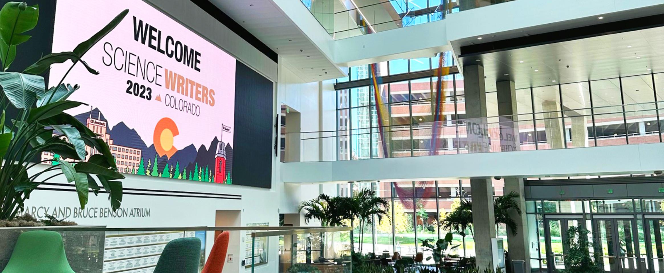 Horizontal photo of the spacious sunlit atrium of the Anschutz Health Sciences Building, with a giant wall screen displaying the Science Writers 2023 logo. Chairs are seen in the foreground, and deeper in the background. Photo by Christina O'Haver.