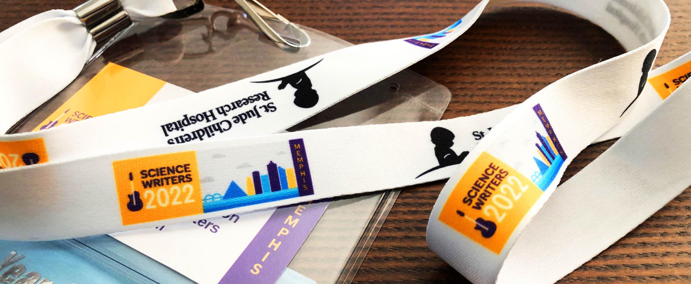 Horizontal photo of a Science Writers 2022 name badge and lanyard draped artistically on a wood-grain table top. The conference logo and St. Jude logo are visible, along with an attendee ribbon attached to the name badge.