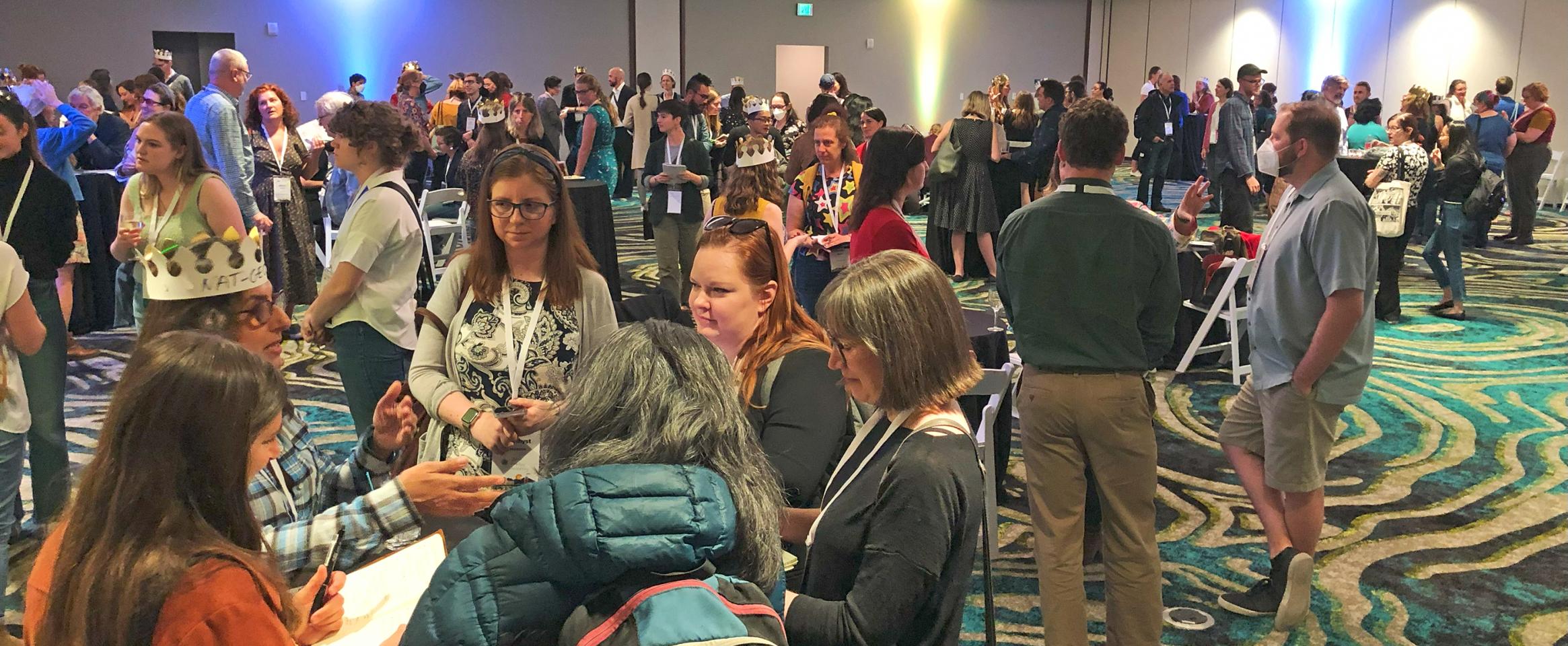 Horizontal photo of a hotel meeting room opened wide with circles of participants gathered around high top tables. Some people are wearing paper crowns. Photo by Ben Young Landis.