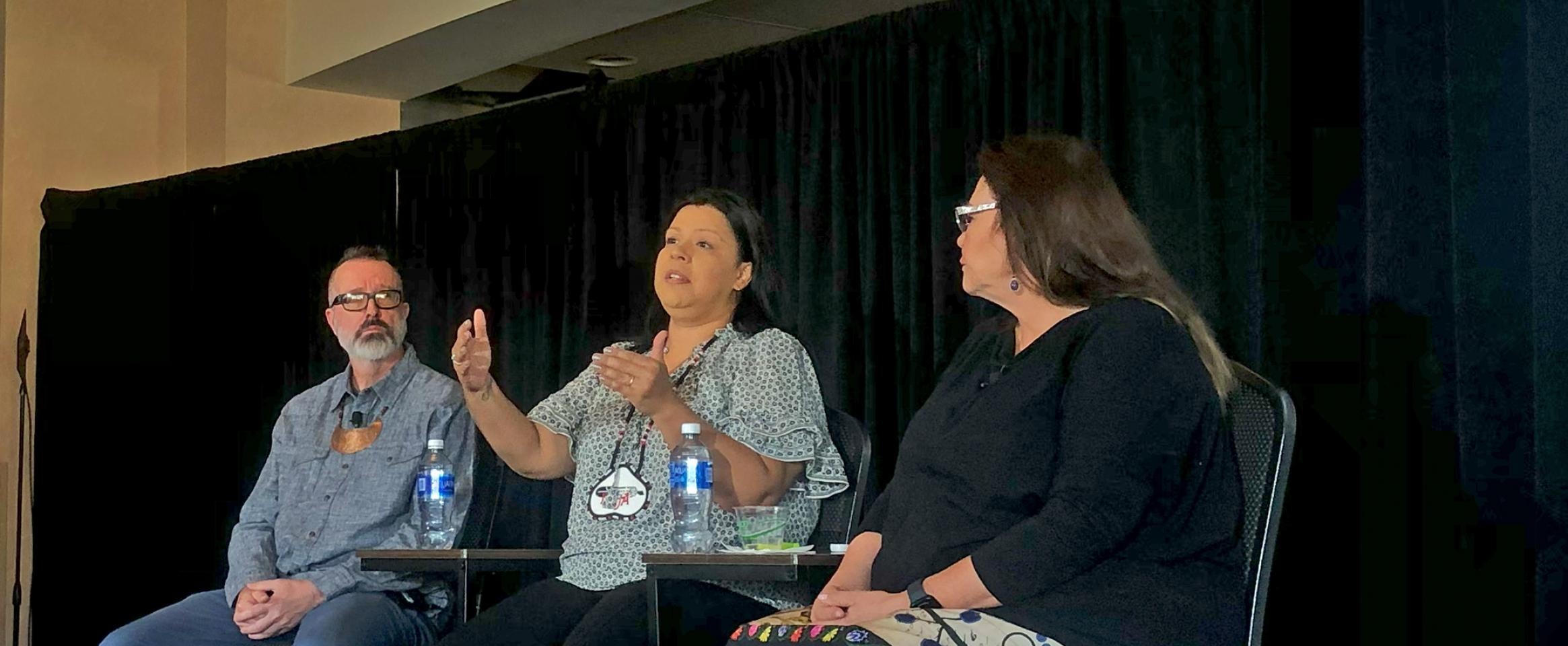 Horizontal photo of Bryan Pollard and Jodi Rave Spotted Bear listening to Francine Compton speak, all sitting alongside one another on stage. Photo by Ben Young Landis