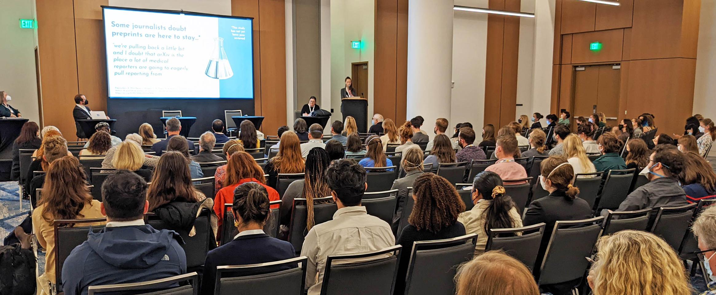 Horizontal photo of the Preprints session at Sci Wri 22 shot from back of the room, showing rows of seats filled with attendees. Panelists are seated on the stage, and the screen slide reads Some journalists doubt preprints are here to say. Photo credit David Jen.