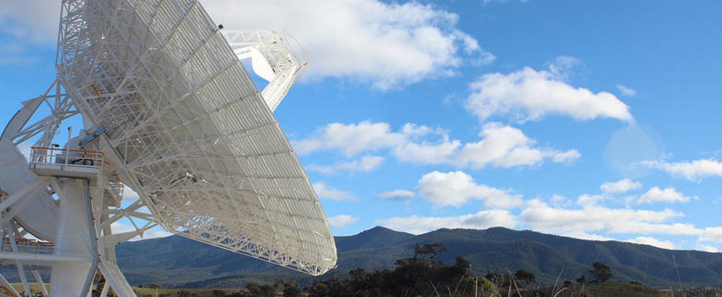 Photo of Deep Space Station 36, a gigantic antenna dish situated against rolling Australian hills and clear sky with clouds.