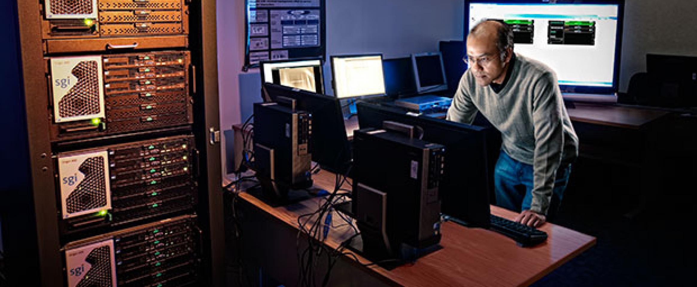 Photograph of a man working in a dimly lit office full of computer server banks and screens.