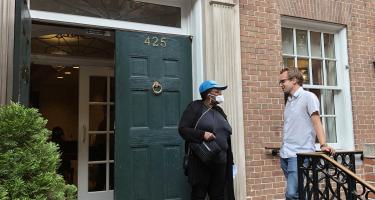 Photo of Neelise, a Fountain House member, talking with Andrew, a Fountain House and N A S W member, on the doorsteps of Fountain House's Manhattan building in New York City. The building has old double doors and a ring knocker, and a stone beam engraved with the organization's name overhead.