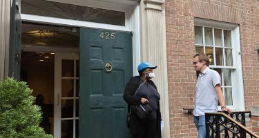 Photo of Neelise, a Fountain House member, talking with Andrew, a Fountain House and N A S W member, on the doorsteps of Fountain House's Manhattan building in New York City. The building has old double doors and a ring knocker, and a stone beam engraved with the organization's name overhead.