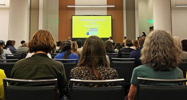 Photo focused on the back of the heads of three audience members amid a sea of attendees in the foreground, while in the background sit panelists David Quarmmen, Sabrina Imbler and Mara Grunbaum below a projector screen with slide title Synecdoche and Science Writing, with moderator Maya L. Kapoor standing at the podium to stage left.