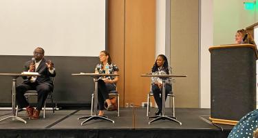 Horizontal photo of Jermey N. A. Matthews, Luba Ostashevsky, and LLathea Mondesir, seated on stage, and moderator Lina Zeldovich, standing at podium.