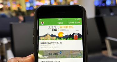 Horizontal photo of a melanated hand holding up a smart phone showing the Science Writers 2023 Whova conference homepage, In the background, airport seating and people are visible. Photo by Paul Adepoju