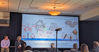 Photo shows conference attendees and panelists Nick Messenger and Stephanie Czekalinski looking at a slide with an illustrated figure showing the multiple chemical exposure pathways that occurred immediately following the disaster and during cleanup operations. Photo by Helina Selemon