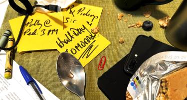 Horizontal photo of a placemat full of sticky notes for family appointments, crayons, mugs, a dirty spoon, a research paper, cereal crumbs, and a blueberry. Photo by Ben Young Landis.