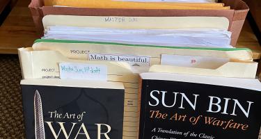 Rectangular photo of Dana Mackenzie's source books, including The Art of War and The Art of Warfare, and file folders of papers.