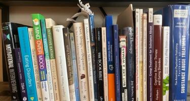Rectangular photo of Claire Wilcox’s office bookshelf showing works on topics including emotional eating, overeating, eating disorders, diet and drugs, substance abuse, self-therapy, and mindfulness. Photo credit: Claire Wilcox.