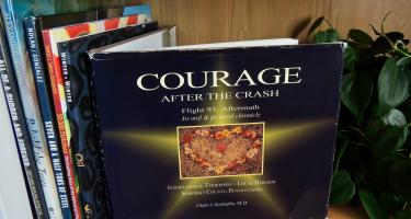 Rectangular photo of Rebecca E. Hirsch’s office bookshelf showing books on terrorism and its aftermath, examples of resilience, such as the restoration of the flag that flew at the World Trade Center on 9/11, and tree-planting projects. Photo credit: Rebecca E. Hirsch.