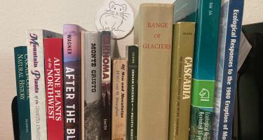 Rectangular photo of David Williams’ office bookshelf showing works on the natural history of the Cascades, the 1980 Mt. St. Helens eruption and ecological responses to it, glaciers, mountain plants, Indian legends, and Tahoma. Photo credit: David Williams.