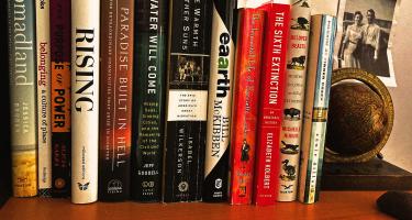 Rectangular photo of a row of books on a wooden bookshelf, with many titles related to the earth, environment, and science. A bookend with an antique globe, and a monochrome vintage photo of a couple are visible adjacent.
