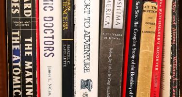 Rectangular photo of a close up view of books on a bookshelf, with spines facing out and many titles related to atomic science and history.