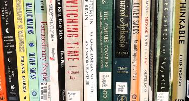 Horizontal photo of a bookshelf with various book spines visible, many with library catalog stickers, and a few authored by Oliver Sacks. Photo by Marc Dingman