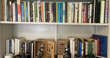 Rectangular photo of Erin Zimmerman’s office bookshelf showing works on botany, trees, naturalists, evolution, ontogeny and phylogeny, Darwin, and evolution, along with two antique student microscopes. Photo credit: Erin Zimmerman.