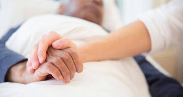 An older person lies in a hospital bed. A younger person holds his hand.