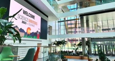 Horizontal photo of the spacious sunlit atrium of the Anschutz Health Sciences Building, with a giant wall screen displaying the Science Writers 2023 logo. Chairs are seen in the foreground, and deeper in the background. Photo by Christina O'Haver.