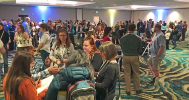 Horizontal photo of a hotel meeting room opened wide with circles of participants gathered around high top tables. Some people are wearing paper crowns. Photo by Ben Young Landis.