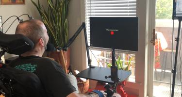 A man in a wheelchair is using assistive technology to interact with a computer screen. He is facing a monitor that displays the question "What do you feel like?" above a red square. He is wearing a black shirt and has a microphone positioned near his mouth, along with a joystick and switch control device secured to his hand. The setup is in a well-lit room with a large potted plant, a window with blinds, and a sliding glass door that opens to a balcony with outdoor furniture and a colorful blanket.