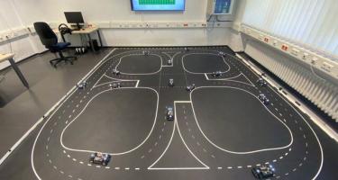 Dr. Bassam Alrifaee’s indoor lab setup at RWTH Aachen University, showing a room with a racetrack-like walled platform on the floor.