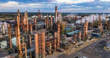 An aerial photograph of the sprawling Pine Bend Oil Refinery in Rosemont, Minnesota, at sunset. Smoke is rising from structures in the background.