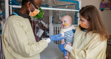 Two gowned researchers who built a personalized CRISPR therapy for a life-threatening liver condition hold an infant boy who was the first to be treated.