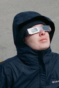 Photo of Kat Escner wearing a hooded shell coat and eclipse sunglasses looking up at the sky