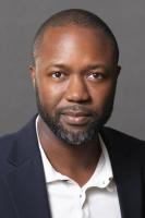 Vertical headshot of Ashley Smart with close cropped hair, beard, suit jacket and studio backgdrop